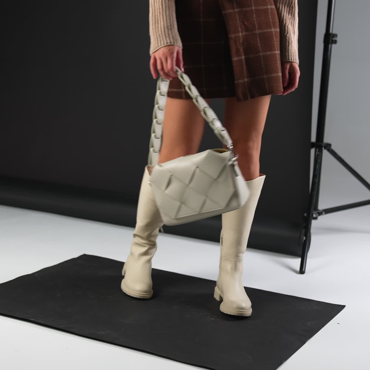 Girl in short skirt and sweater shows light high boots and same color bag. Footwear and accessories demonstration in studio