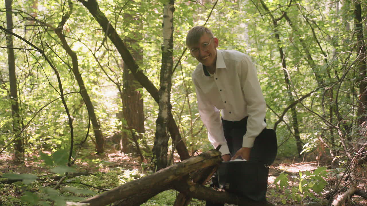 Man in white shirt putting white coat inside backpack bag while standing in forest, surrounded by green trees and sunlight, preparing for activity with calm and focused movement