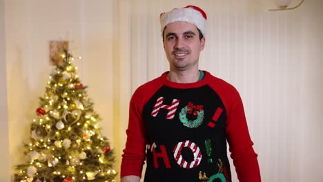 happy young man with a ho-ho-ho sweater and santa hat having fun in front of a christmas tree and pointing with a finger at the camera