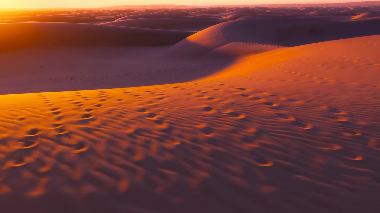 Dune dorate del deserto si estendono attraverso l'inquadratura mentre il sole tramonta, proiettando una luce arancione e lunghe ombre.
