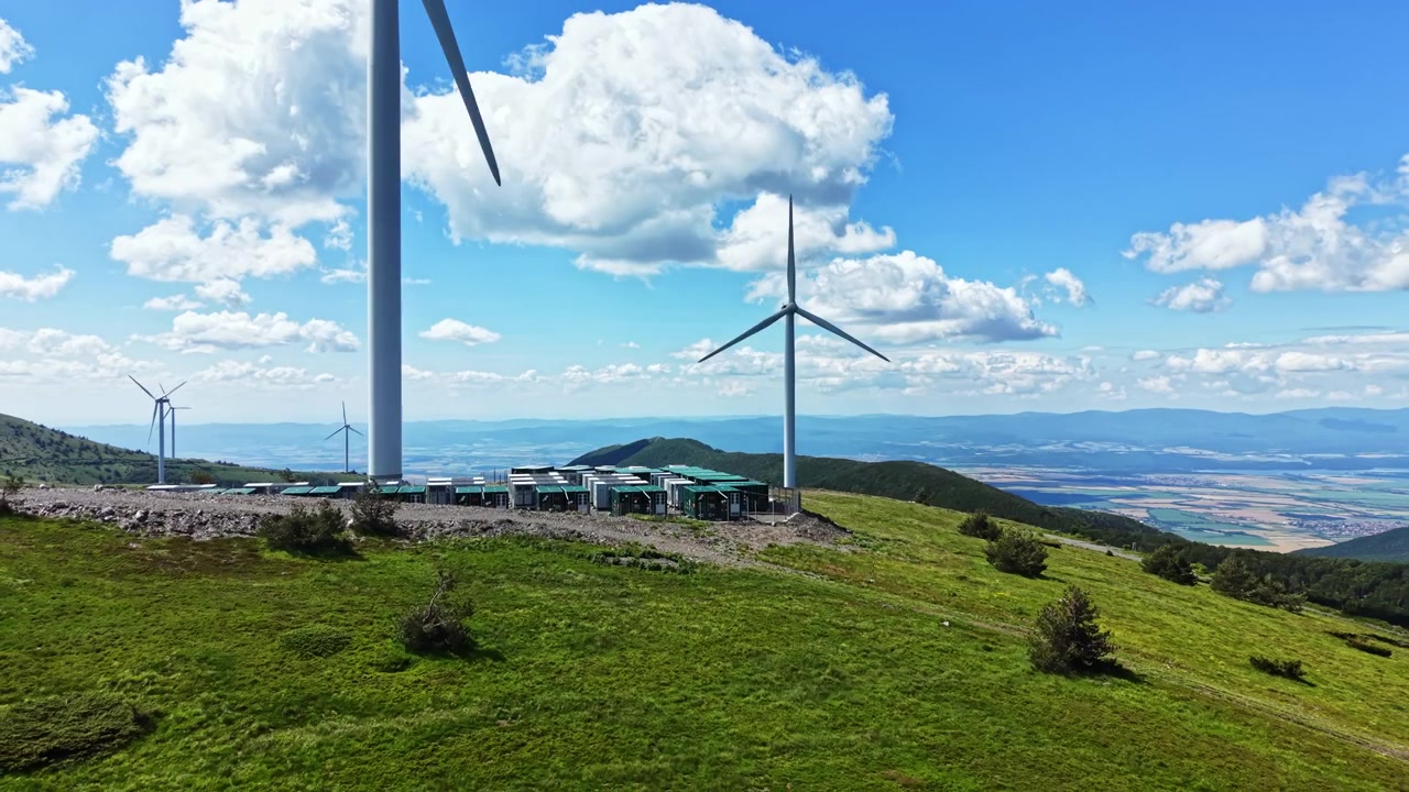Turbine eoliche si ergono alte su una collina verde, fornendo energia pulita in una zona rurale sotto un cielo azzurro brillante con nuvole sparse.