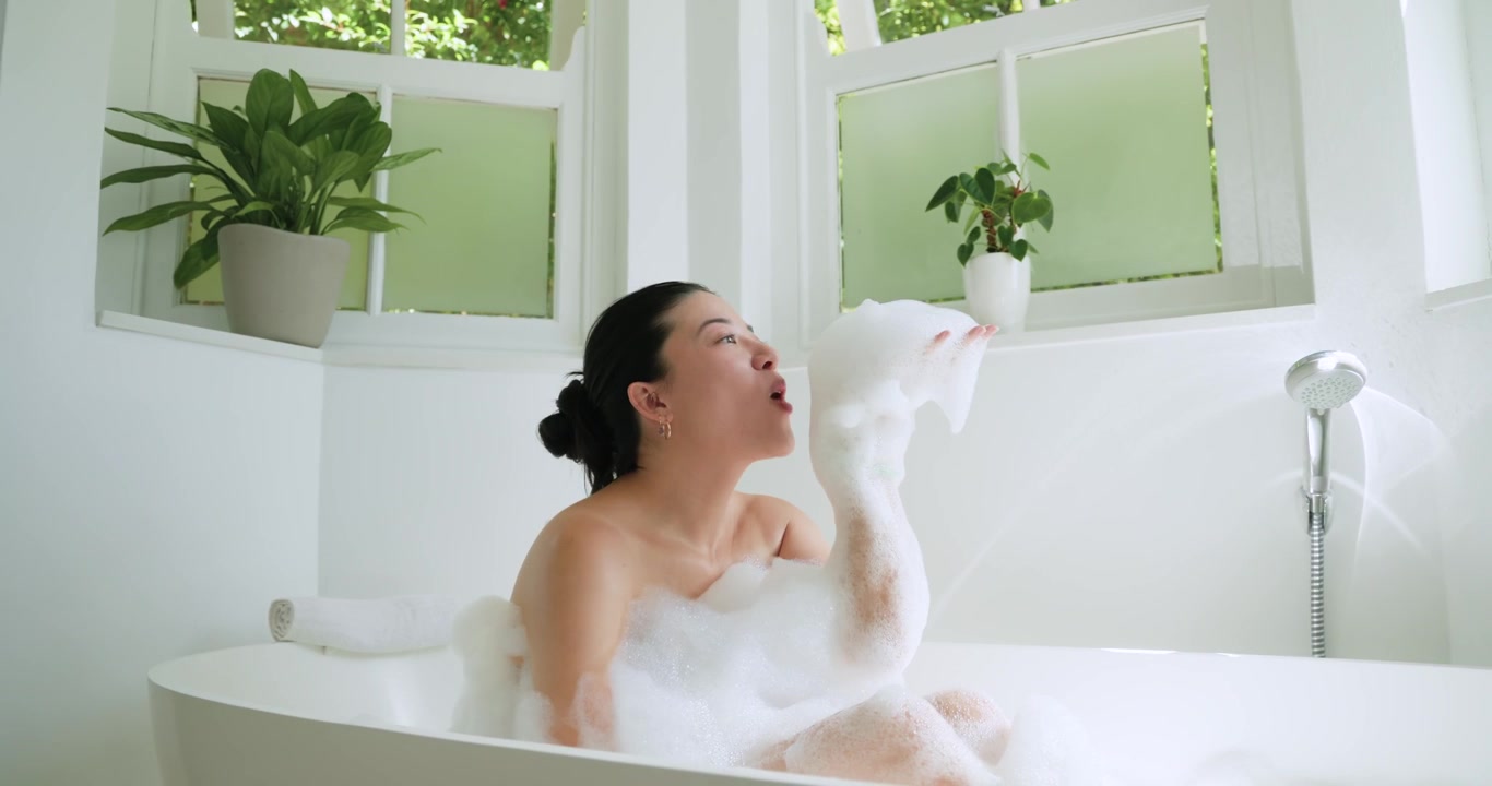 Donna afroamericana che si muove, raccoglie bolle e lancia schiuma nella vasca da bagno a casa, rilassandosi.