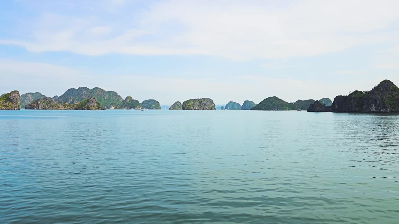 The tranquil waters of Ha Long Bay stretch toward the horizon, where countless limestone karst formations create a dramatic silhouette against the soft blue sky, reflecting the bay's legendary beauty