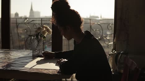 Female-artist-is-sitting-by-the-table-with-flowers-on-it,-drawing-on-paper-with-pen,-in-a-very-minimalistic-art-studio-on-the-background.