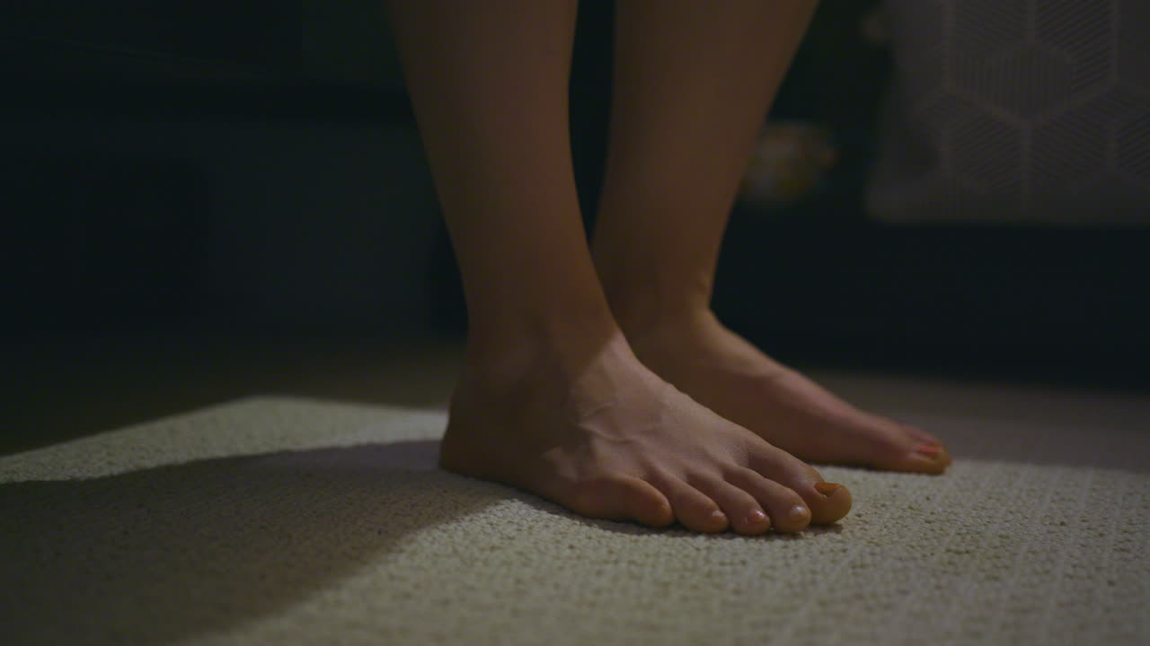 Primo piano dei piedi di una donna mentre si alza lentamente dal letto ed esce dall'inquadratura.