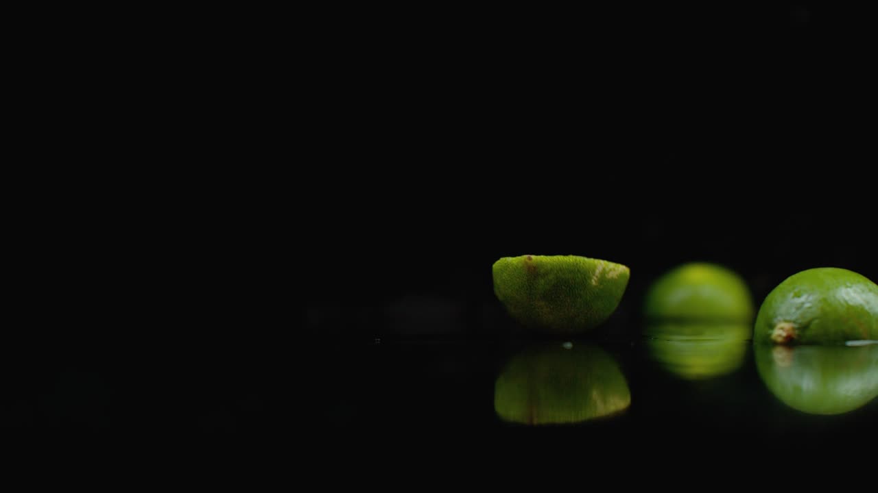 dos limas maduras caen sobre un vaso con salpicaduras de agua en cámara lenta sobre un fondo oscuro.