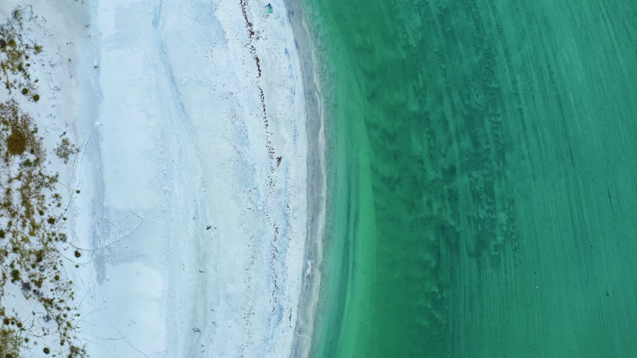 Turquoise water curls along a pale sand shoreline as shifting textures beneath the surface create a serene coastal scene filled with quiet motion and natural rhythm