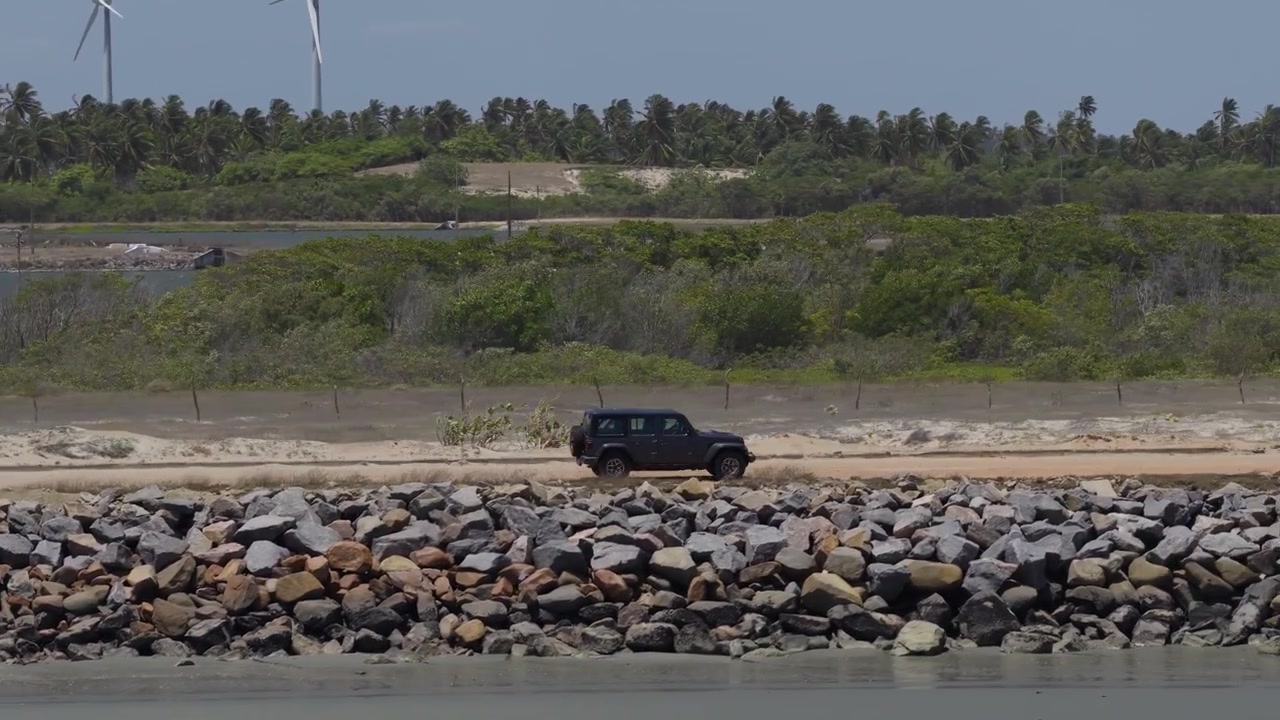 Una carrellata professionale riprende un SUV scuro che passa davanti a infrastrutture industriali e rigogliosi palmeti. L'imponente scala di una turbina eolica definisce il paesaggio costiero del Ceará, Brasile.
