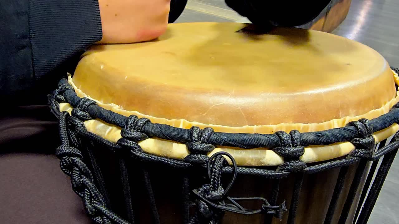 A musician is playing with his bare hands a djembe bongo drum, tapping their hands on the surface to create a rhythm. The camera moves around to capture different angles of their performance.