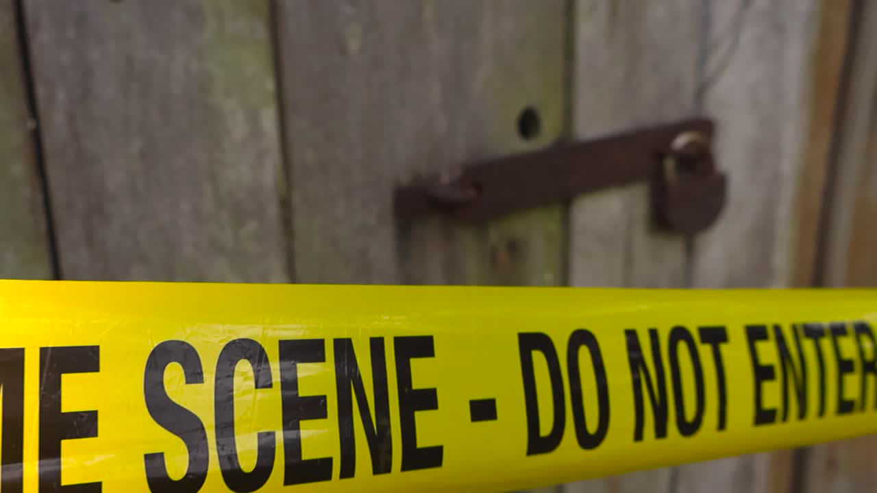 Close up view gliding along a yellow colored police investigation forensic crime scene tape that is in front of a wooden barn or farm house door, with a rusty padlock holding the wooden door shut