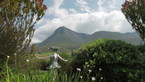 Estatua-De-Niña-En-El-Jardín-De-Vegetación-Nativa,-Con-Vistas-A-Hermosas-Montañas-Con-Nubes-Que-Se-Avecinan