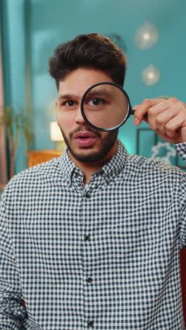 indian young man holds magnifying glass near face looking at camera with big zoomed eye analyzing