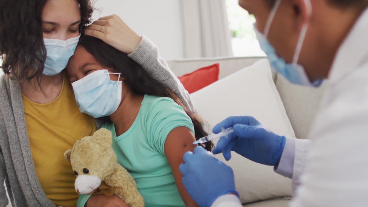 Hispanic male doctor making injection to girl with mother at home, all wearing face masks,