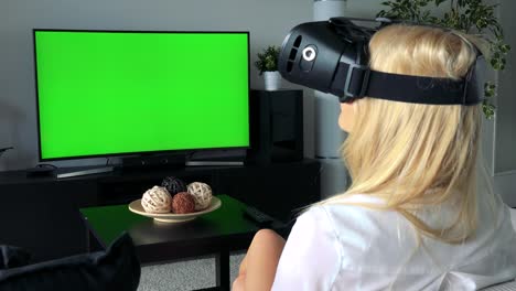 young attractive blond woman uses virtual reality glasses in home, in living room - green screen television in background
