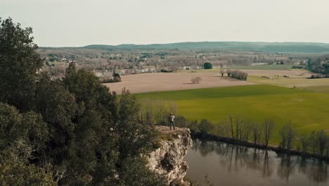 Very-wide-shot-behind-a-forest-of-a-woman-on-a-cliff-taking-nature-photos,-wide-aerial-view-with-the-river-in-s-in-the-background