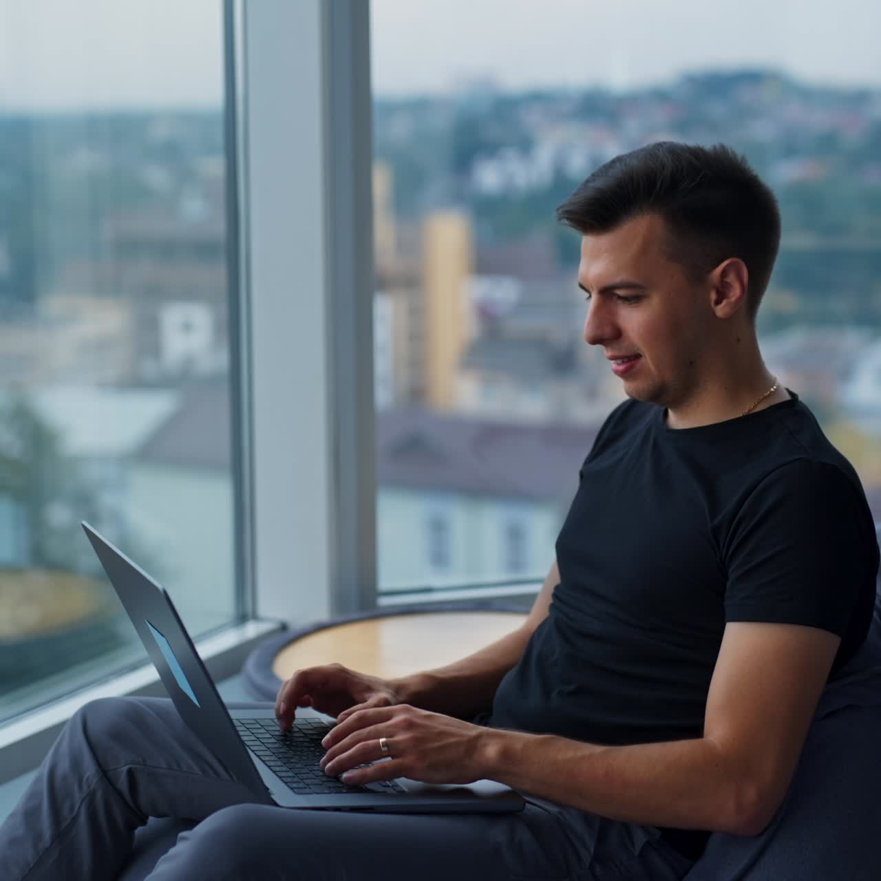 Young creative handsome freelancer working at computer. Male self-employed entrepreneur working sitting in bean bag chair near window. Blurred backdrop