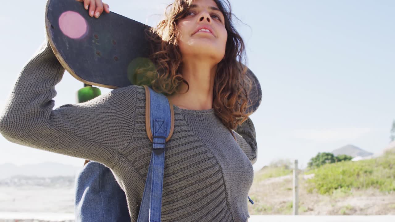 mujer de raza mixta con patineta detrás de la cabeza de pie en el sol disfrutando de la vista por el mar