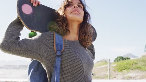 Mujer-De-Raza-Mixta-Sosteniendo-Patineta-Detrás-De-La-Cabeza-Parada-Al-Sol-Disfrutando-De-La-Vista-Junto-Al-Mar