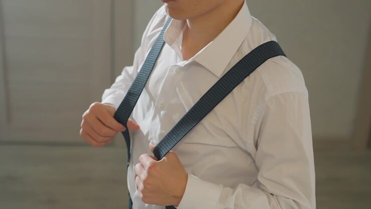 Asian man checking suspenders before leaving, confident posture and composed expression, hands smoothing shirt collar, crisp white collar and dark pinstripe trousers with clips, soft shadowing