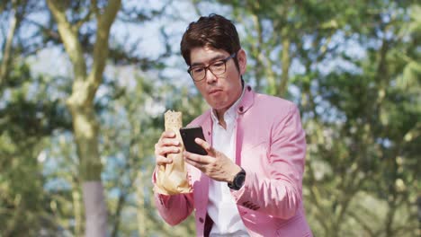 Asiatischer-Mann,-Der-Einen-Snack-Isst-Und-Sein-Smartphone-Benutzt,-Während-Er-Im-Park-Sitzt