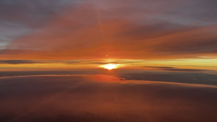 Un'immersiva visuale in prima persona dalla cabina di pilotaggio di un jet, durante un volo al tramonto nell'alta atmosfera, tra strati eterei di nuvole color arancio.