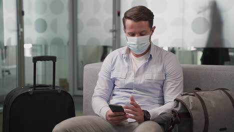 a young man wearing a mask consults his cell phone while waiting for his flight to depart in an airport lounge.