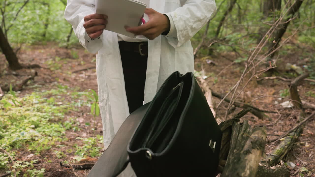 Close view of biologist in white lab coat with black backpack on old tree stump reaching to take out jotter and pen in natural forest environment surrounded by greenery and blurred background