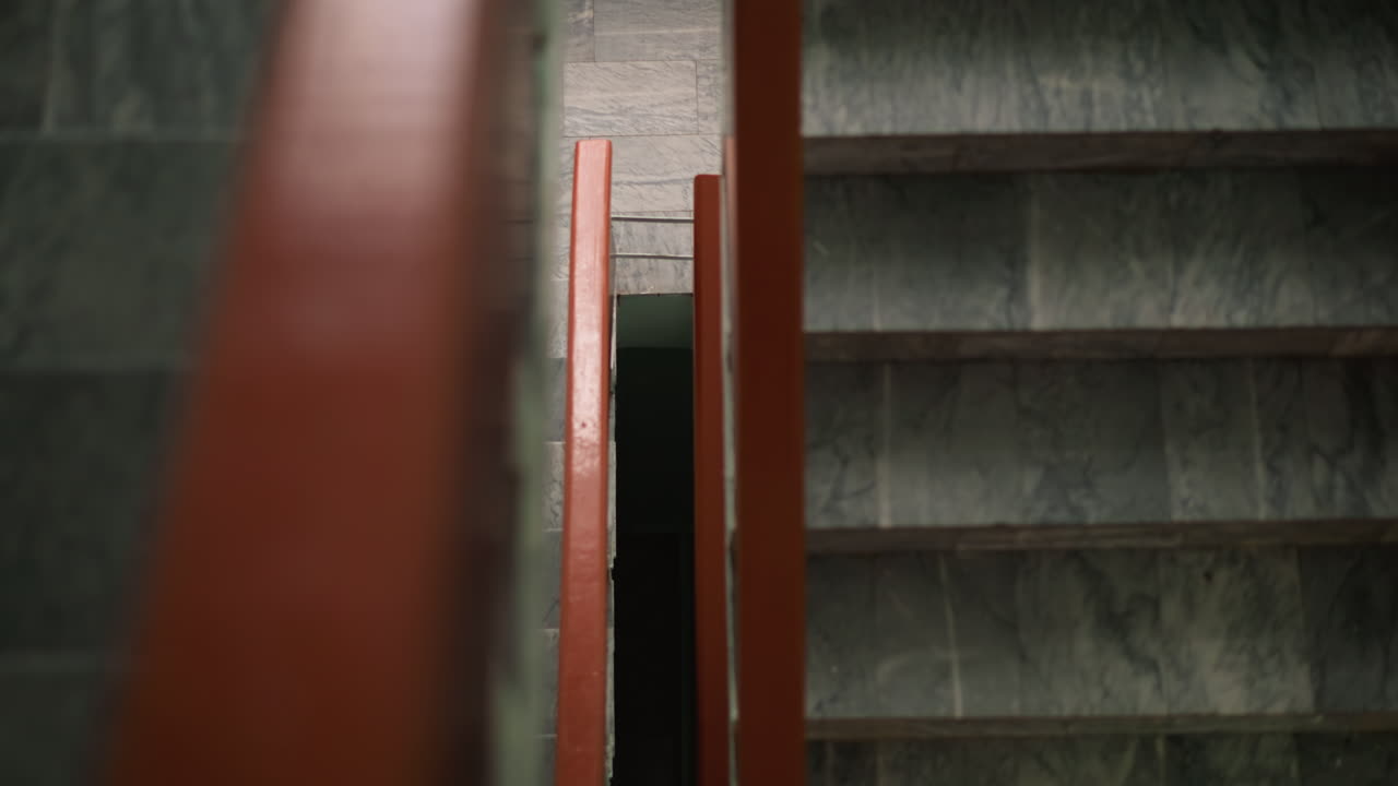 Close view of staircase with red handrail and marble steps, view of woman in white top and black leggings walking upward, moody corridor, soft focus framing suggesting quiet movement and minimalism