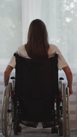 brunette woman moves wheelchair closer to bright window thinking about life. person with disability suffers from loneliness after leg injury backside view