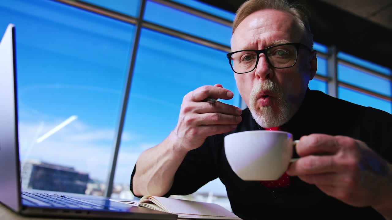 Fair-haired bearded senior man wearing glasses holding a white cup. Man blows on the drink and waves hand with a pen in it. Low angle view.