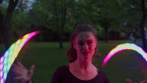 Smiling-Woman-Twirling-LED-Fans-in-Twilight-Lit-Park,-Medium-Shot