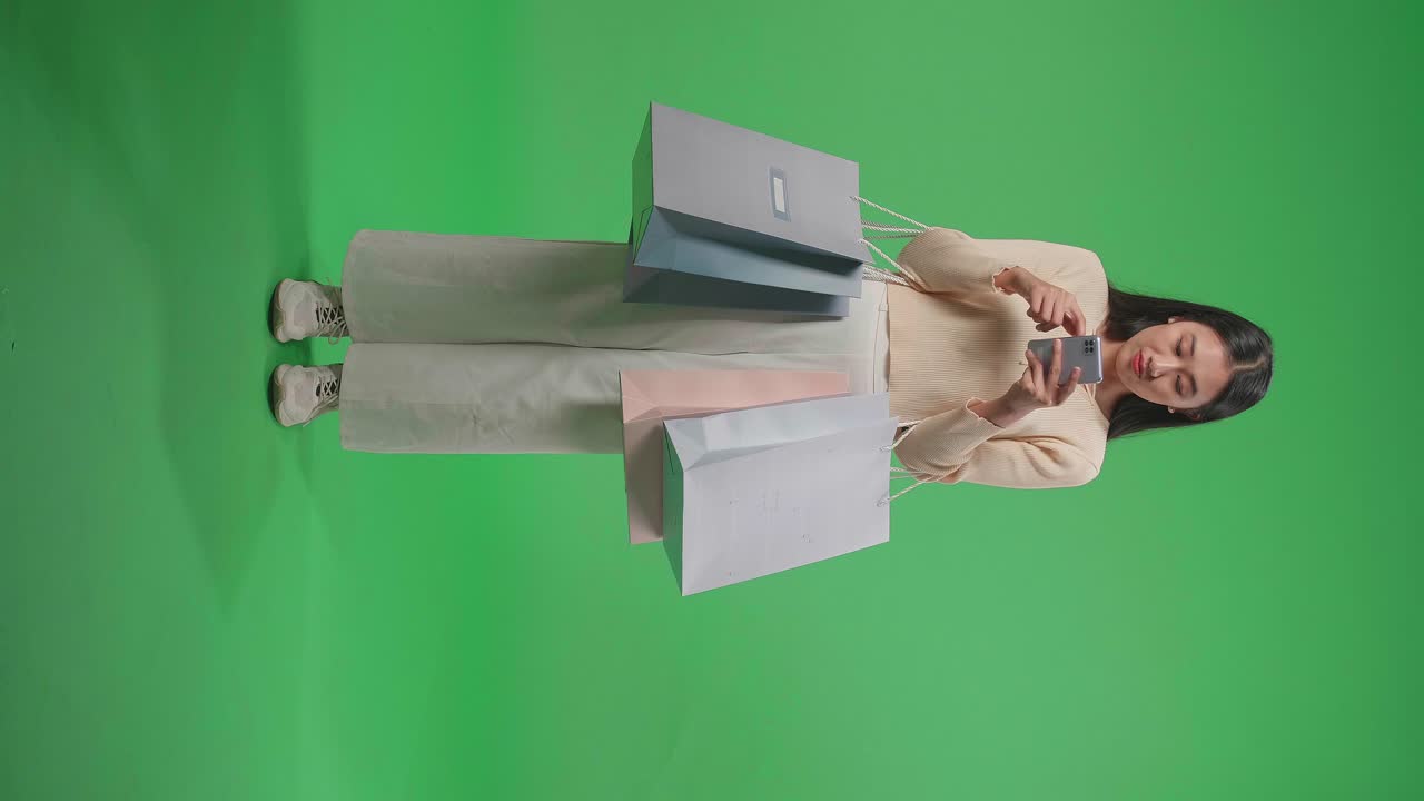vista completa de una mujer de compras asiática con bolsas de compras sosteniendo un teléfono móvil antes de señalar y sonreír a la cámara mientras está de pie frente a la pantalla verde