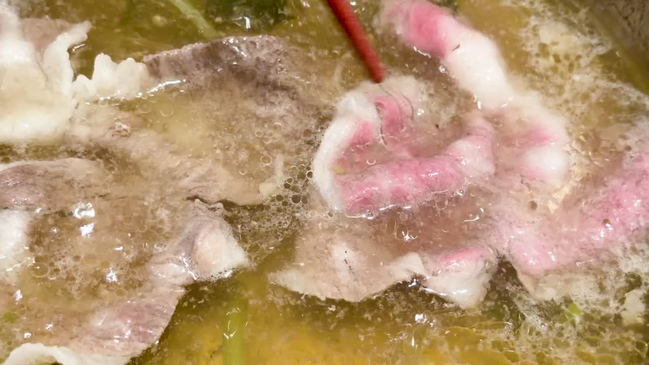 Chopsticks cook marbled wagyu beef slices in boiling broth, close-up, overhead, warm lighting