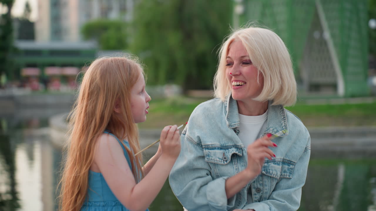 close up riverside scene mom with red manicure laughs as daughter playfully touches moms face with paint brush while holding painting board during warm sunset art bonding moment in urban park setting