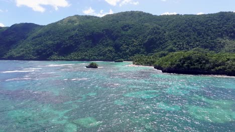 slow aerial flight over clear water with coral reef and greened mountains in background