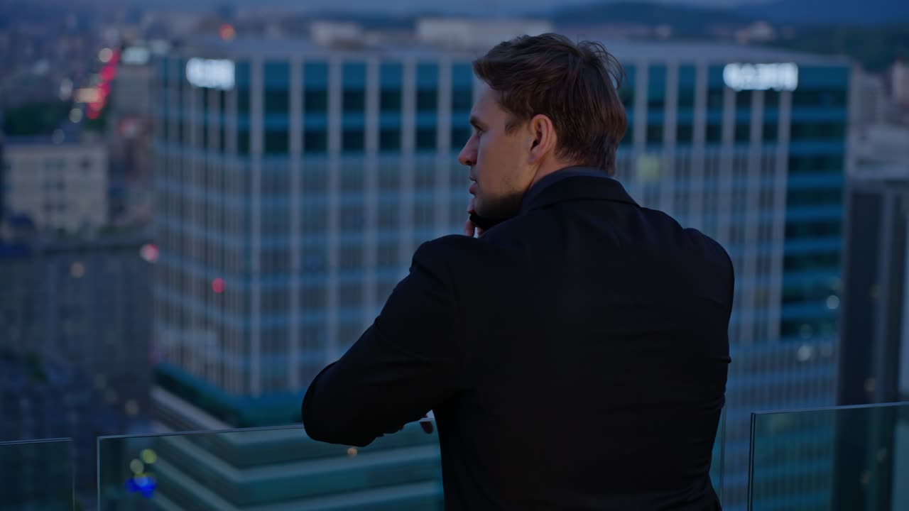 Rear view of businessman in suit talking on phone on rooftop terrace overlooking modern city skyline at dusk