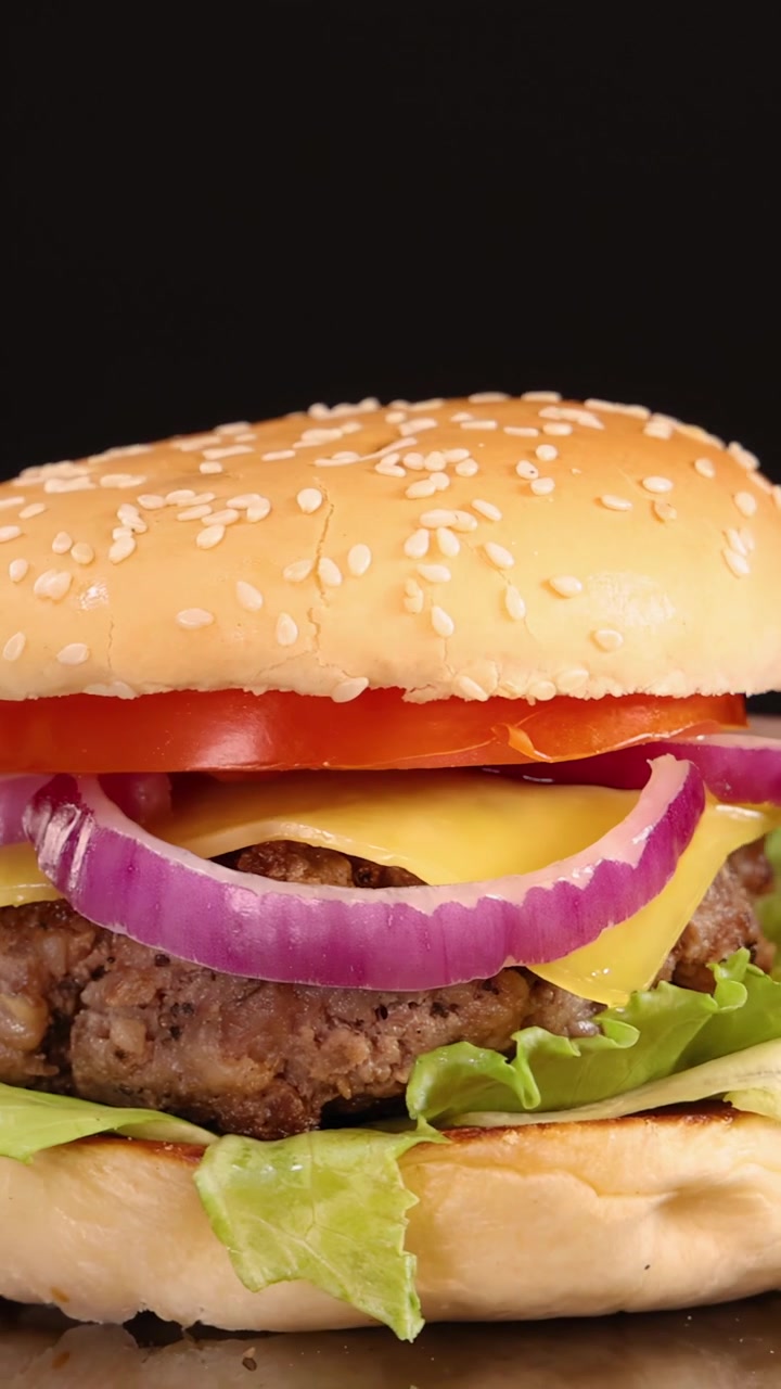 Un succoso hamburger di manzo con formaggio, lattuga, pomodoro e cipolla ruota fluidamente sotto una brillante illuminazione da studio, isolato su uno sfondo nero senza soluzione di continuità.