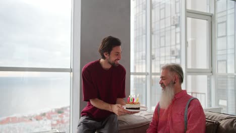 un hombre moreno con barba en una camiseta roja trae un pastel y velas de cumpleaños encendidas para su padre en la vejez con cabello gris y una barba exuberante en una camisa rosa en un apartamento moderno con vistas al mar