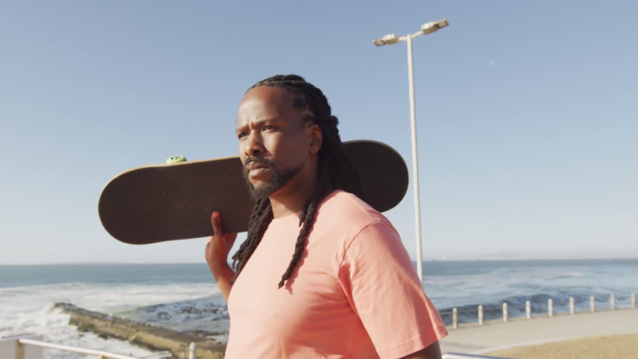Video af afroamerikansk mand, der holder skateboard ved havet.