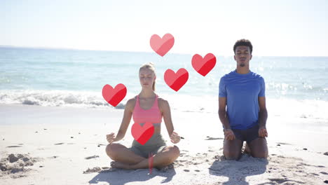 animation of red hearts over diverse couple meditating on beach