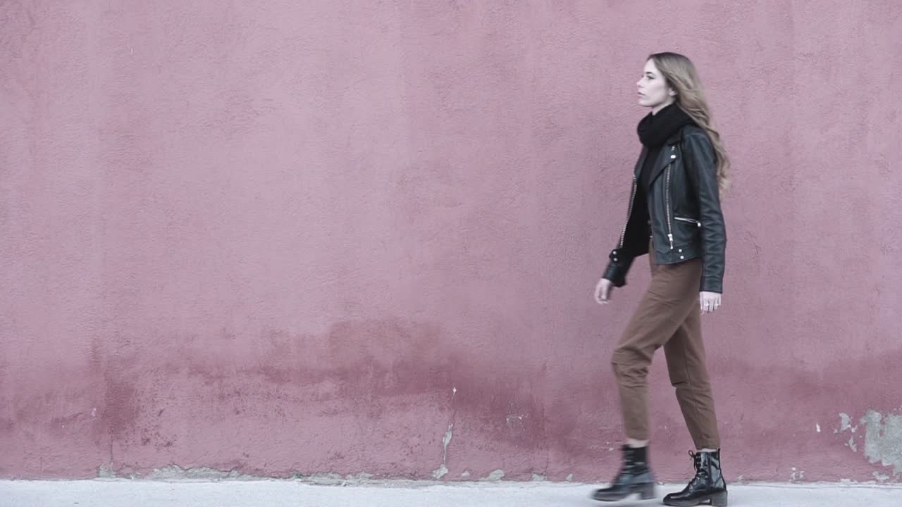 Video al rallentatore di una giovane modella millennial che appare molto sicura di sé mentre cammina in avanti, indossando moderni stivali neri e una giacca di pelle per un look deciso.