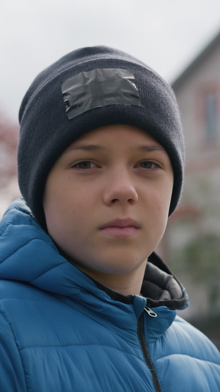 primer plano de un niño con gorra negra y chaqueta azul de pie al aire libre, cerrando y abriendo los ojos pensativamente, con una casa borrosa de techo rojo, follaje de otoño y un entorno al aire libre sereno