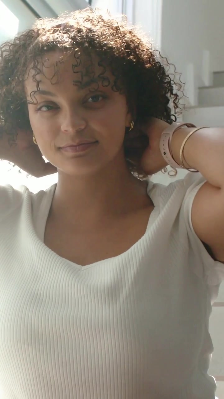 Video vertical: Luz solar iluminando a una mujer tocándose los rizos cerca de la barandilla de la escalera, mostrando un reloj inteligente, cuello en V.