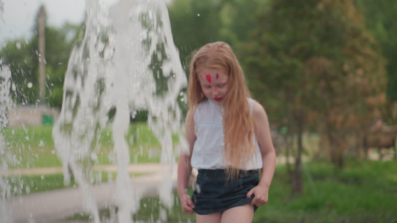 Face painted young girl with head bowed playing near water, boy beside touching fountain, blurred playground behind, summer green setting showing quiet focus and shared exploration of splash