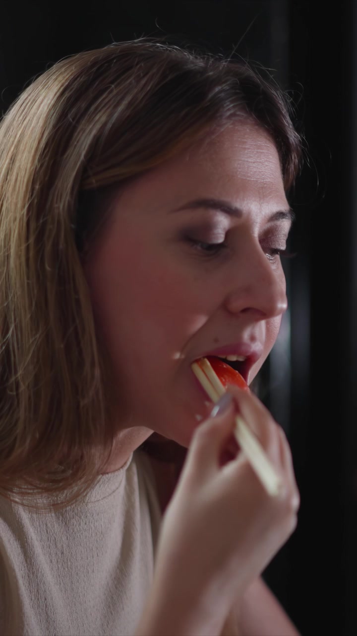Primer plano de mujer comiendo con palillos en la mano. La mujer sostiene y lleva un trozo a la boca, centrándose en su perfil contra un fondo oscuro. La iluminación suave resalta los detalles de la textura del cabello.