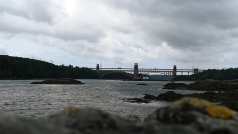 Mirando-Por-Encima-Del-Primer-Plano-Rocoso-Desenfocado-Al-Estrecho-De-Menai-Puente-Britania-A-Anglesey