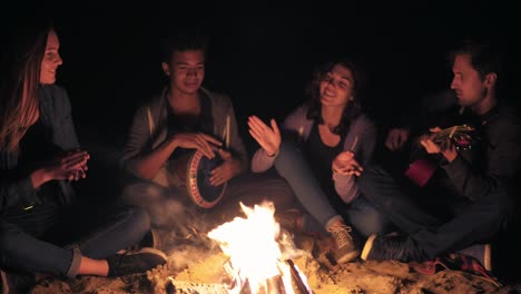 Multiracial-group-of-young-women-and-men-sitting-by-the-bonfire-late-at-night-and-singing-songs,-playing-guitar-and-percussion