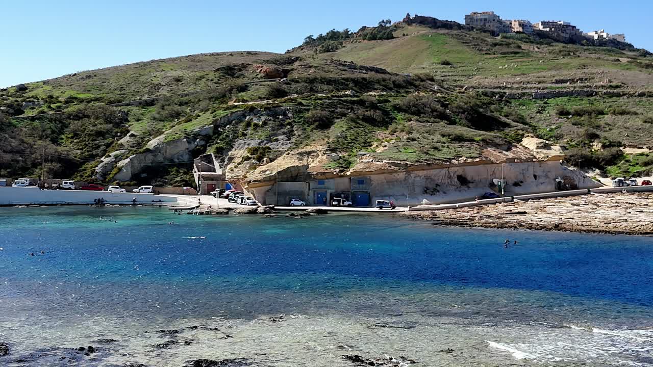 Gozo'daki Qbajjar Koyu; Malta'da parlak, güneşli bir yaz gökyüzü altında oyulmuş tekne garajlarına sahip kireçtaşı kayalıkları, zümrüt yeşili yamaçları ve kristal berraklığındaki turkuaz suları sergiliyor