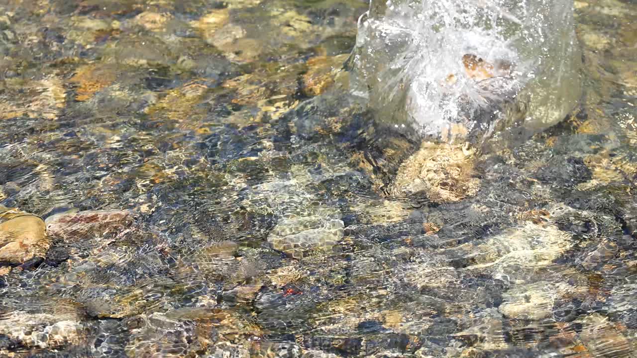 Uma pedra é jogada em um riacho de águas cristalinas, espirrando água sobre pedras coloridas à luz do dia