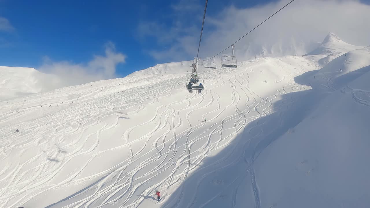 Two-Man Chairlift First-Person View Ascending on Sunny Blue Sky Day with Ski Tracks in Powder and Skiers Below